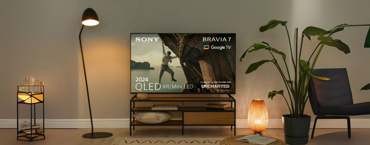 A Sony Bravia 7 QLED TV displayed in a modern living room setup with warm lighting, featuring a floor lamp, wooden TV stand, potted plants, and a comfortable chair. The TV screen showcases a movie scene with the text "2024 QLED XR | Mini LED" and "Google 