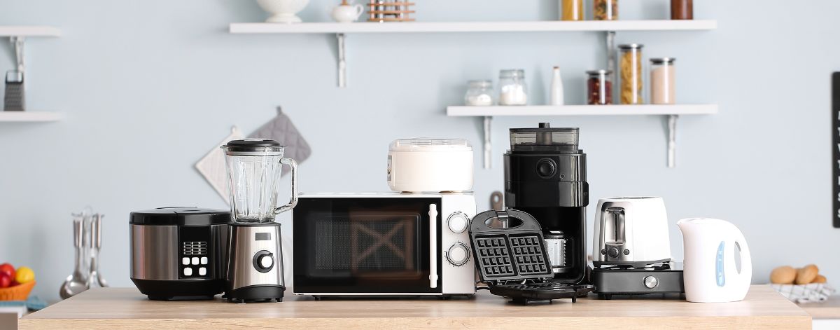  A variety of small kitchen appliances displayed on a wooden countertop, including a blender, microwave, waffle maker, coffee maker, rice cooker, and electric kettle, set against a light blue kitchen background with shelves holding jars and decor.