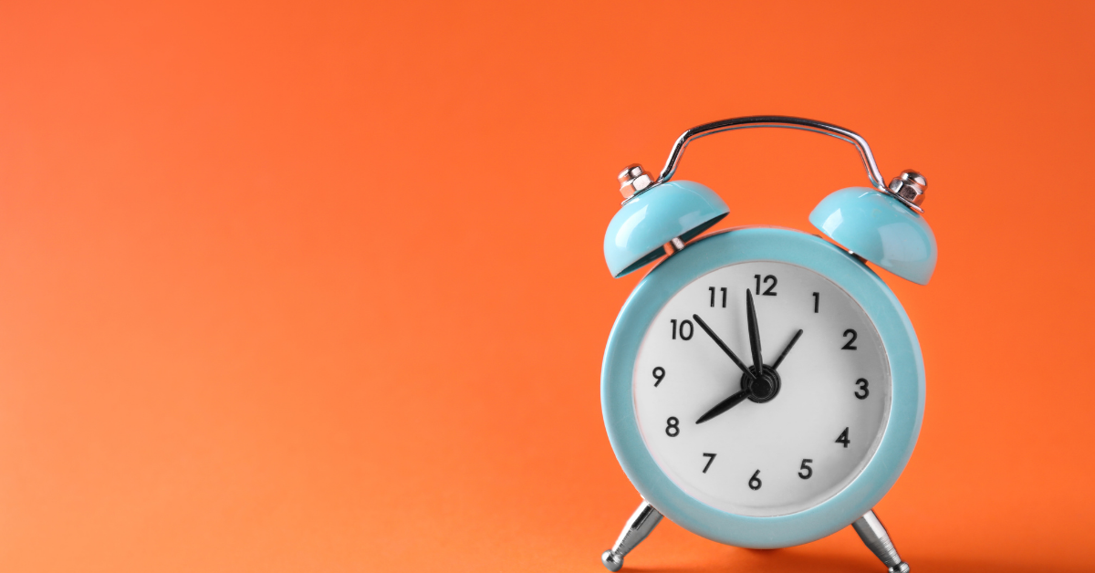 A light blue retro-style alarm clock with black hands and numbers, placed against a vibrant orange background, symbolising the importance of using appliances during off-peak hours to save money on energy bills