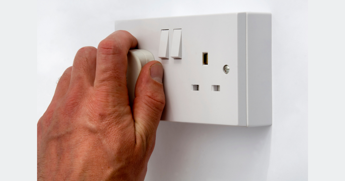 A hand unplugging a power cord from a white wall socket, emphasising the importance of saving energy and reducing electrical usage when devices are not in use